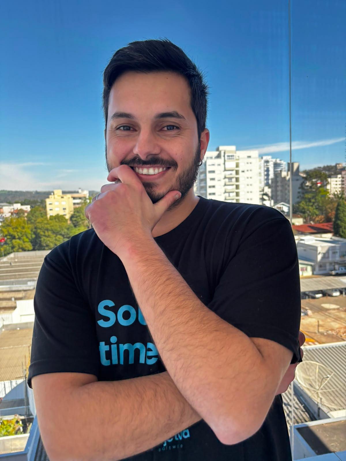 Matheus Maydana smiling, hand resting on his chin, on a balcony with a city view behind him.